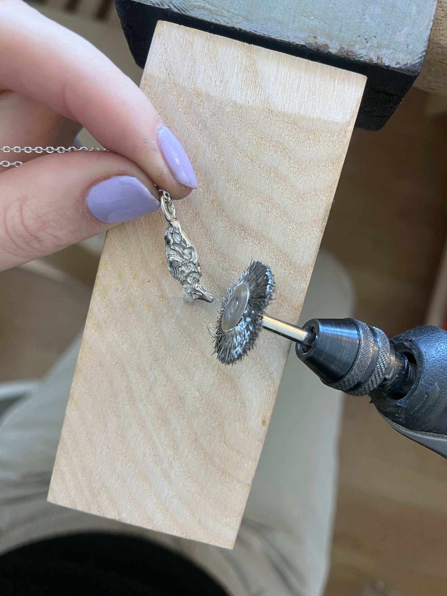Person using a drill to work on a silver earring on a wooden block.